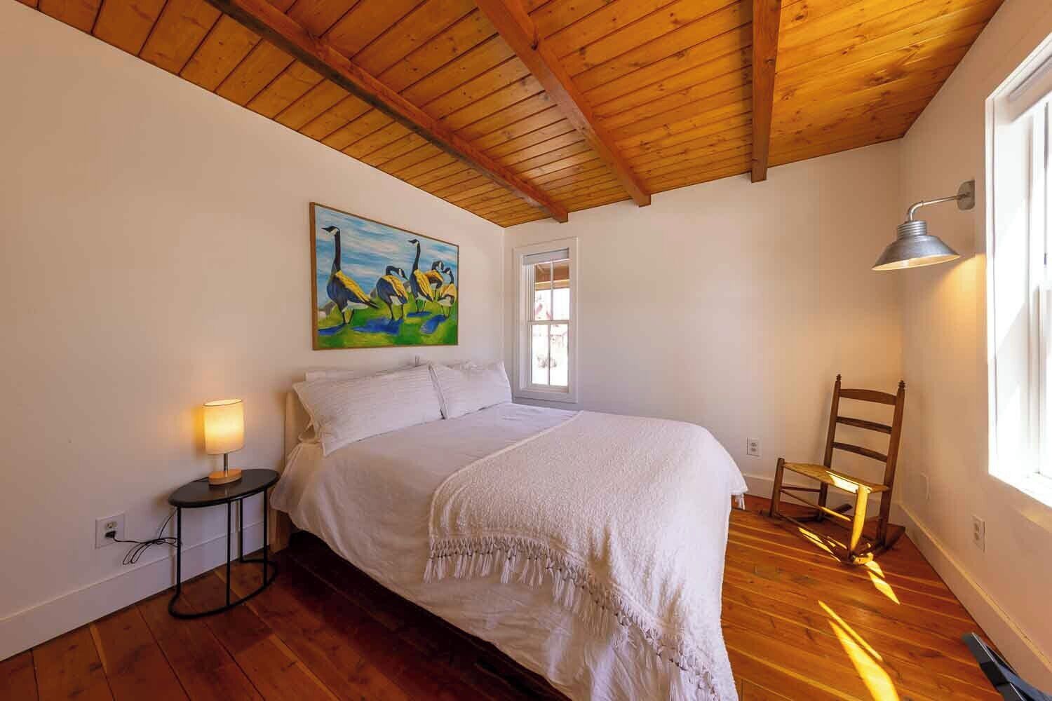 A bedroom with a wooden ceiling, a bed with white bedding, a painting on the wall, a bedside table with a lamp, and a chair near the window.