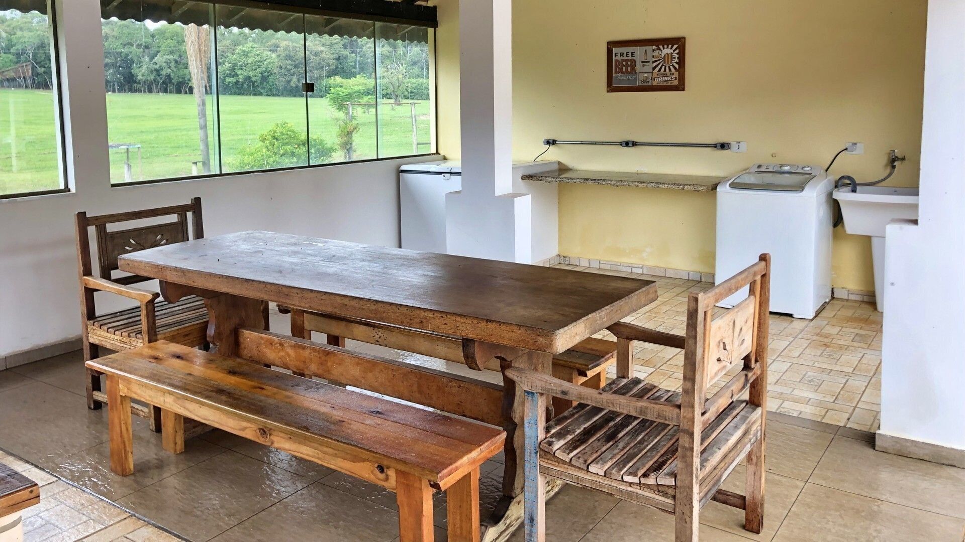 Un tavolo da pranzo in legno rustico con panche, vista su un giardino attraverso ampie finestre, e un angolo cottura con lavello e un cartello che offre birra e bevande gratuite.