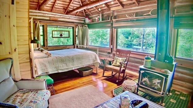 A cozy cabin interior with a wooden ceiling, a bed, a rocking chair, a small table, and a fish tank. - Historic Cabin, Peaceful, Waterside, Patio Firepit (Eureka Springs)
