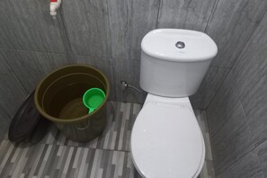 Eine Toilette mit einem grünen Eimer daneben, einem weißen Toilettenpapierhalter und einem gefliesten Boden.
