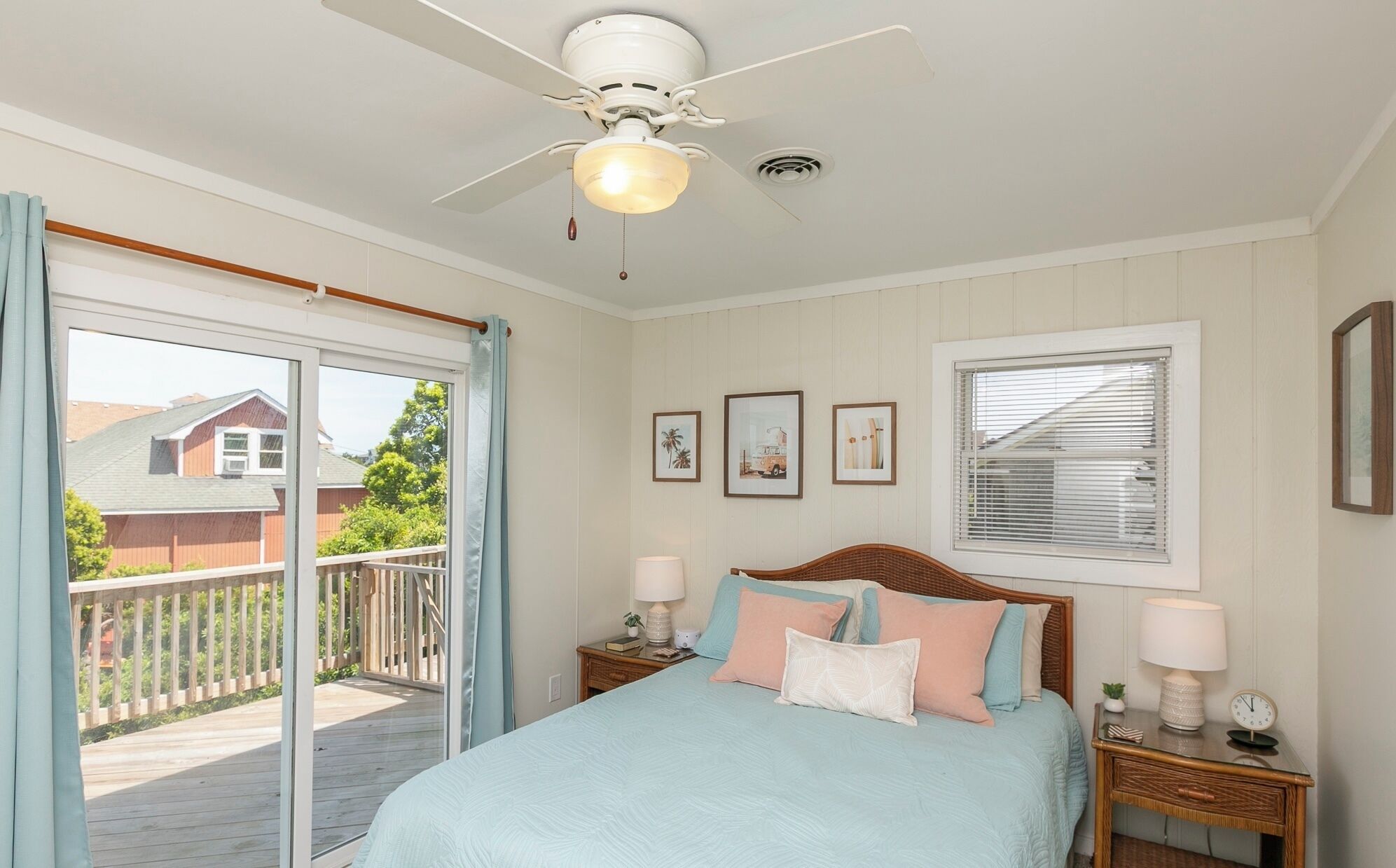 A bedroom with a bed, two bedside tables, a ceiling fan, a window with blinds, and a view of a deck and a house outside.