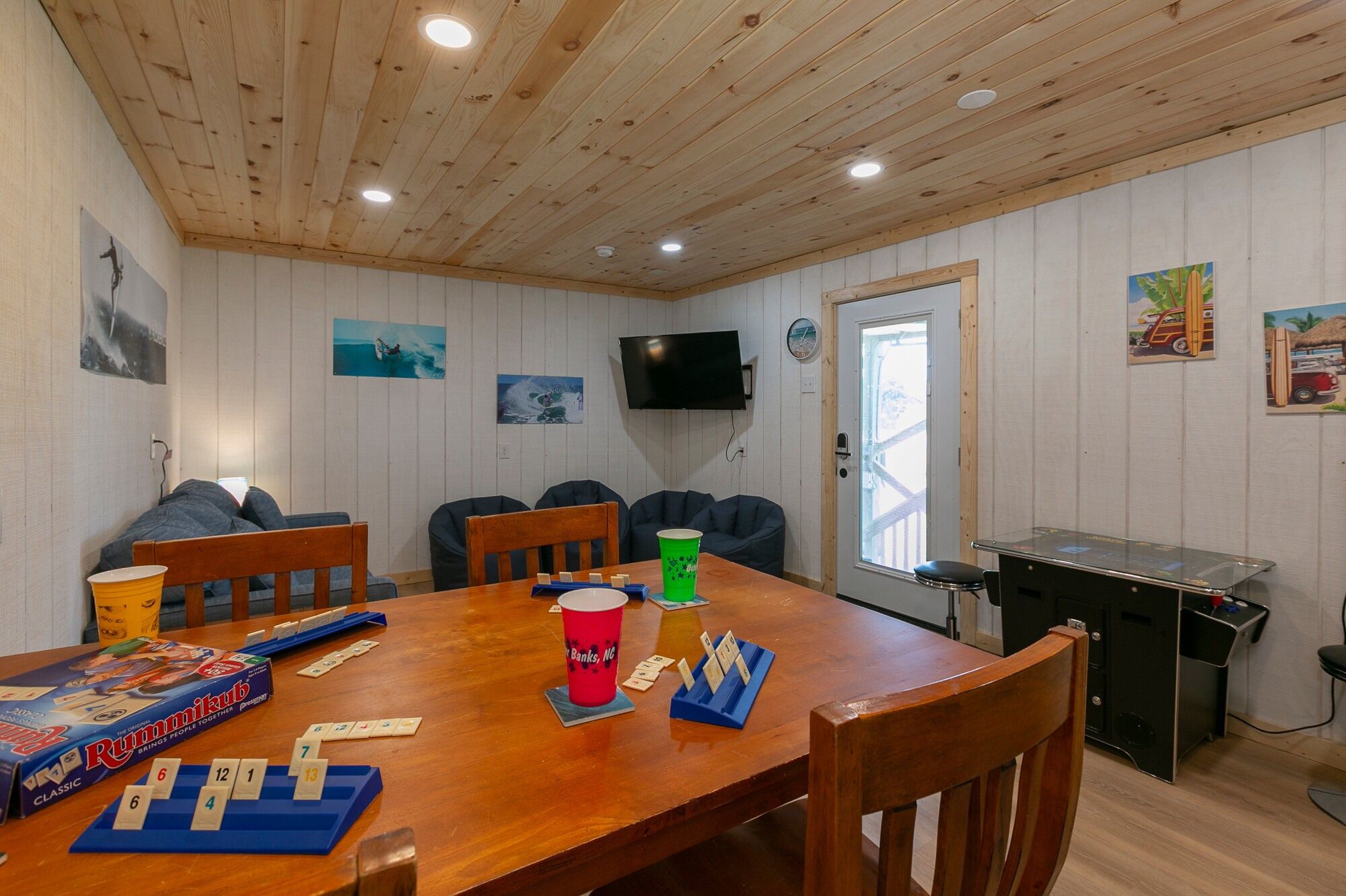 A room with a wooden ceiling, a wooden table with a board game, chairs, a TV, and a window.