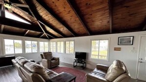 A spacious living room with a high ceiling, large windows, a leather sofa, a coffee table, and a TV. - SunsetSanctuary-CanalFront/DogFriendly/Pool&Tennis (Kill Devil Hills)