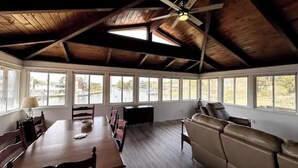 A spacious room with a high ceiling, wooden beams, a dining table with chairs, a sofa, and large windows offering views of the outside. - SunsetSanctuary-CanalFront/DogFriendly/Pool&Tennis (Kill Devil Hills)