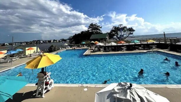 A swimming pool with people swimming and sunbathing, surrounded by umbrellas and lounge chairs. - SunsetSanctuary-CanalFront/DogFriendly/Pool&Tennis (Kill Devil Hills)