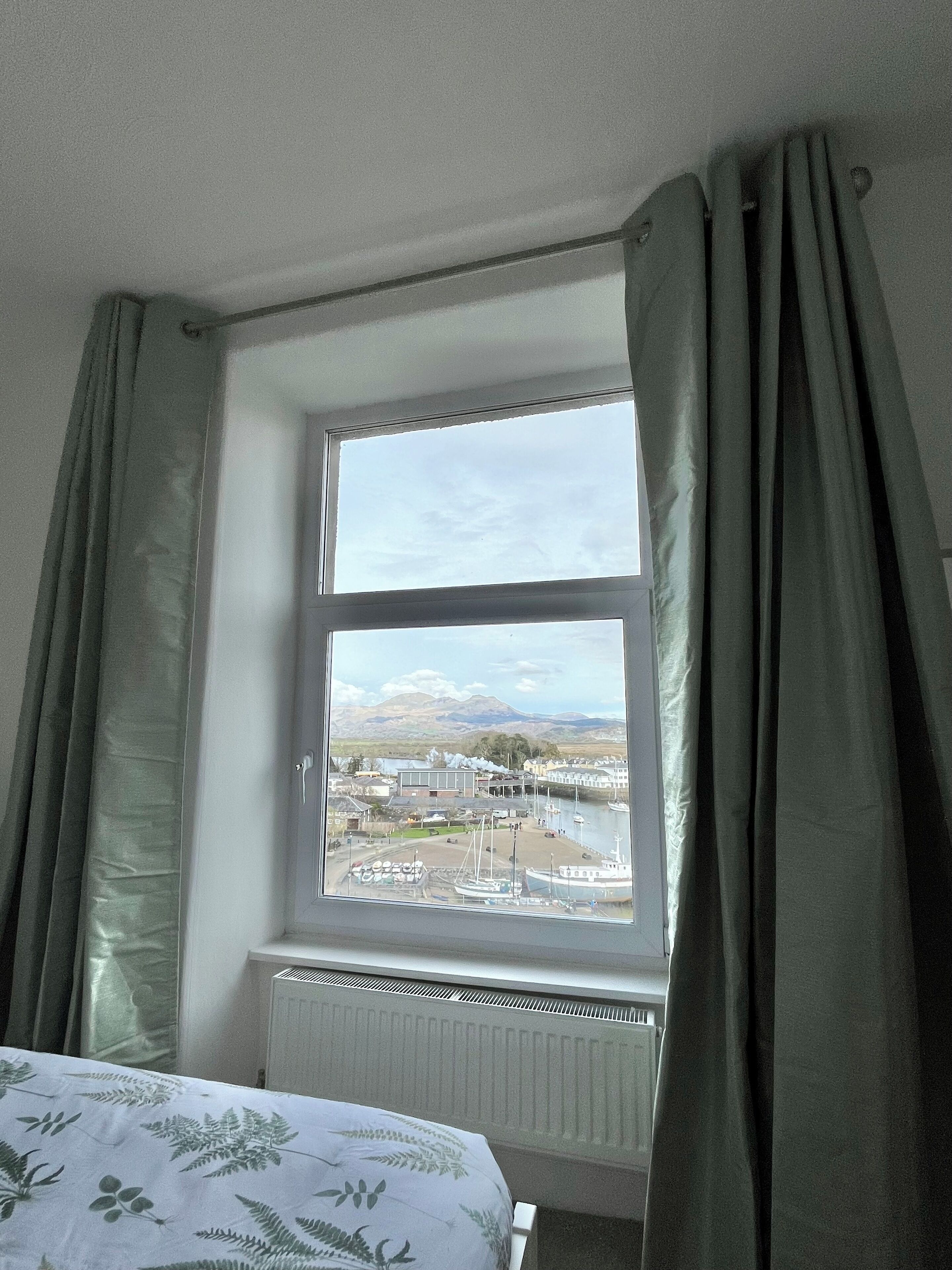 Ein Schlafzimmer mit großem Fenster, Heizkörper und einem Bett mit Blumenmuster.