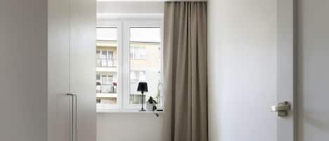 A narrow hallway leading to a bedroom with a bed, a window with curtains, a small stool, and a lamp on a bedside table.