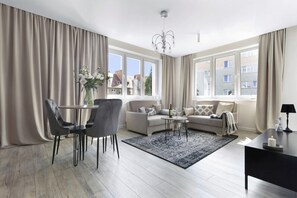 A modern living room with a sofa, dining table, and chairs. There are large windows with curtains, a chandelier, and a view of buildings outside. - Elite Apartments Chicky Deluxe (Gdansk)