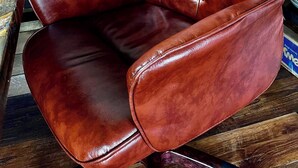 Un fauteuil en cuir marron, doté d’un dossier et d’accoudoirs capitonnés, posé sur un plancher en bois.