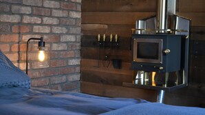 Une pièce avec un mur en briques, un mur recouvert de panneaux de bois, un lit avec une couverture blanche et une cheminée moderne dotée d’une enceinte en verre.