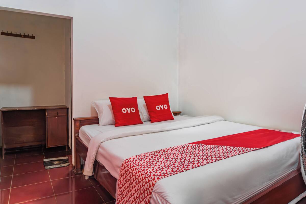 A hotel room with a bed, two red pillows, a wooden nightstand, and a red and white patterned throw blanket.