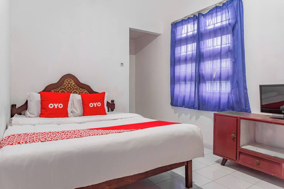 A bedroom with a bed, a red and white bedspread, a red headboard, two red pillows with "OYO" text, a red cabinet, and a blue curtain.