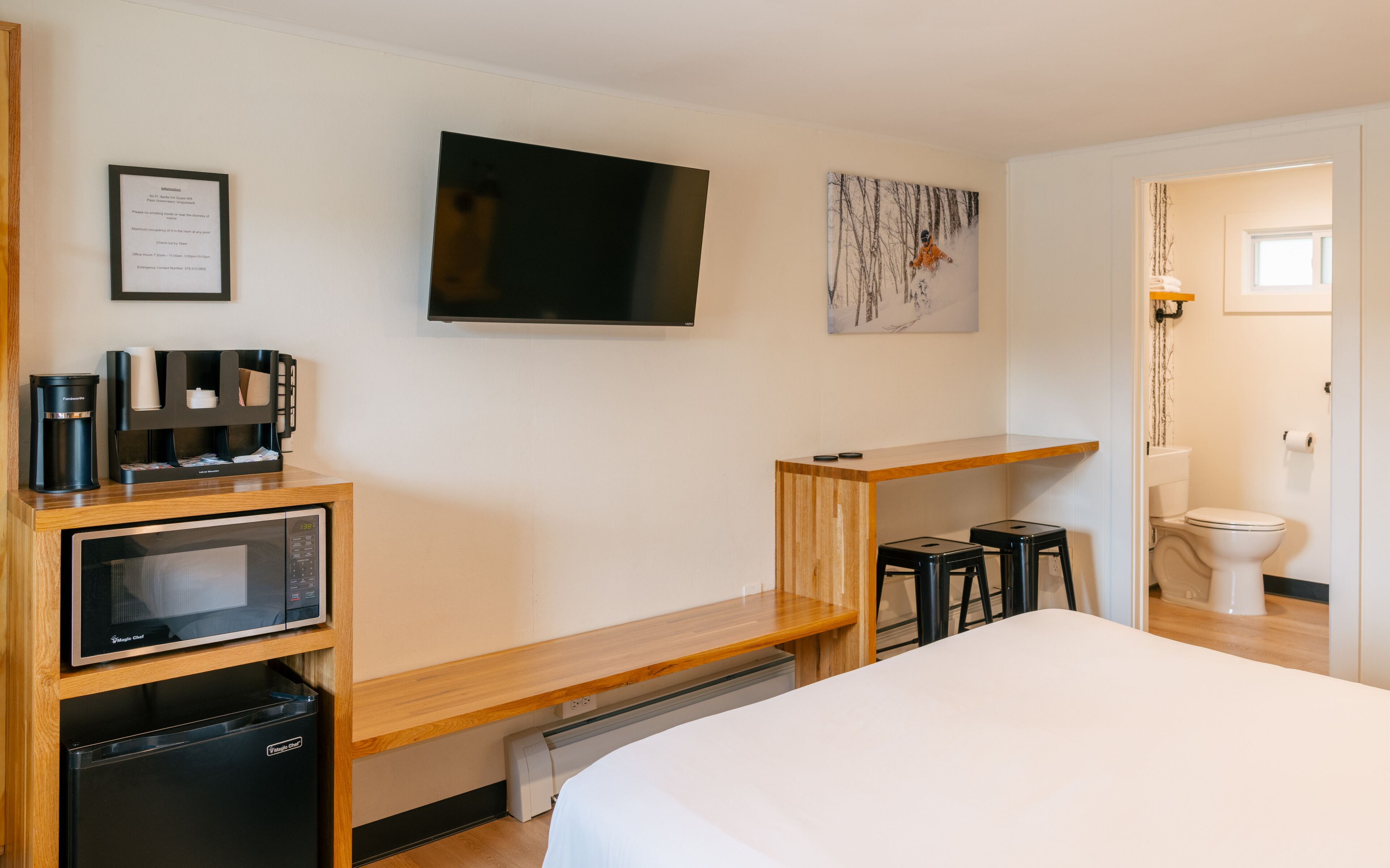 Habitación de hotel con cama, televisión montada en la pared, cafetera, microondas, pequeño escritorio con silla y un inodoro.