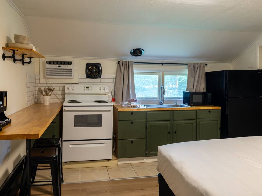 Una kitchenette compacta con estufa, microondas y lavamanos. Hay un taburete pequeño y una cafetera. La habitación tiene armarios verdes y una cama con ropa de cama blanca.