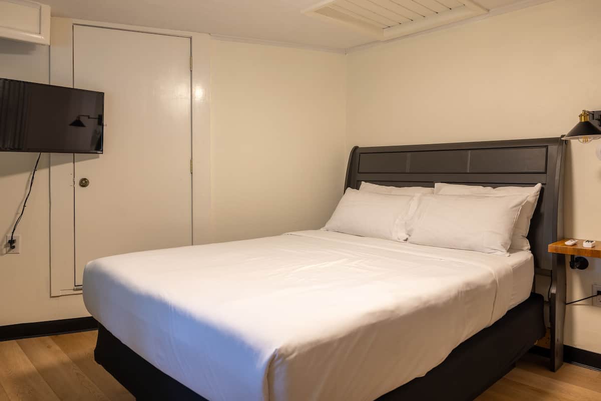 A neatly made bed with white linens in a room with a wooden floor, a flat-screen TV mounted on the wall, and a small wooden nightstand with a lamp.