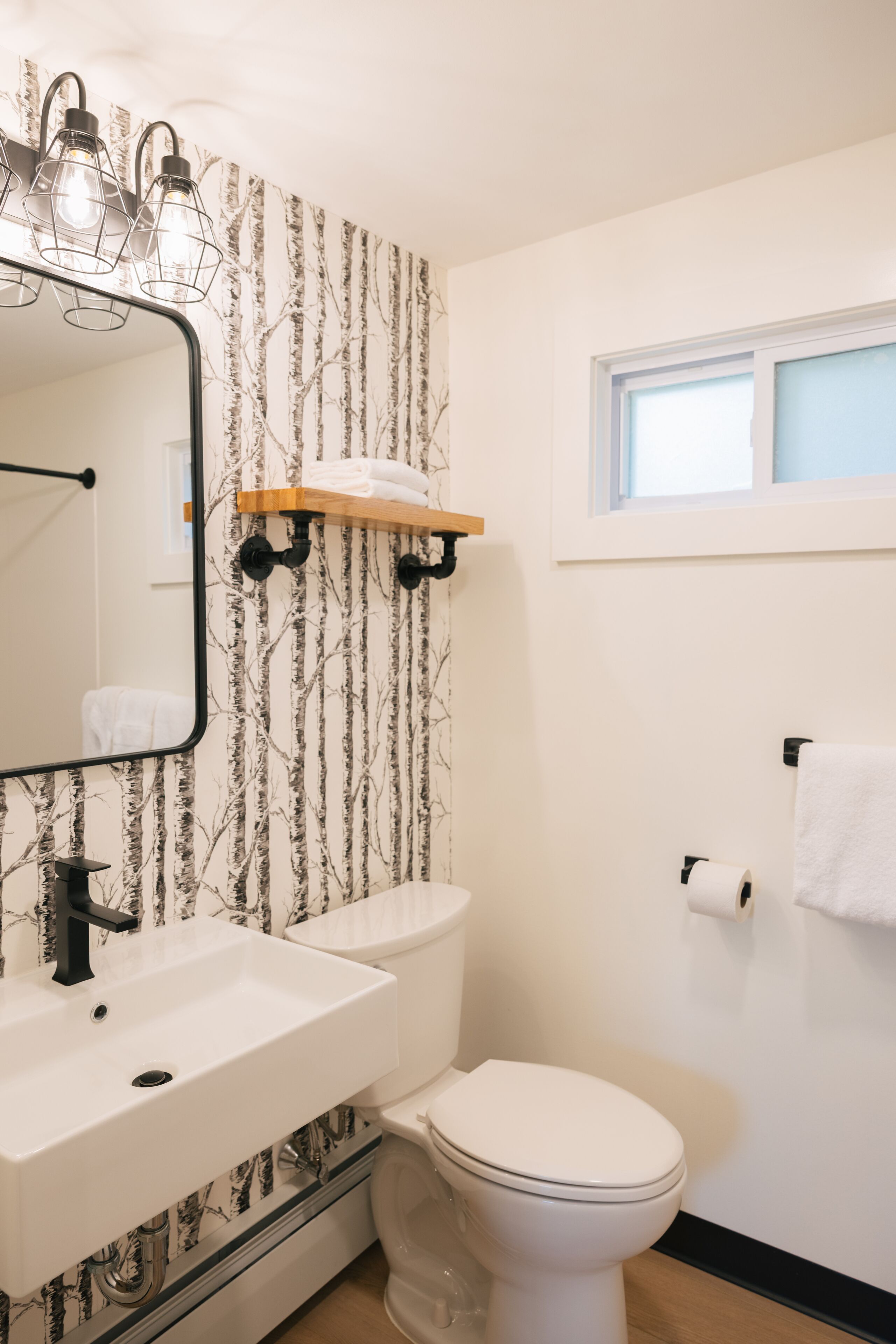 A bathroom with a white sink, a toilet, and a mirror with three lights. There is a window above the toilet and a towel rack on the wall.