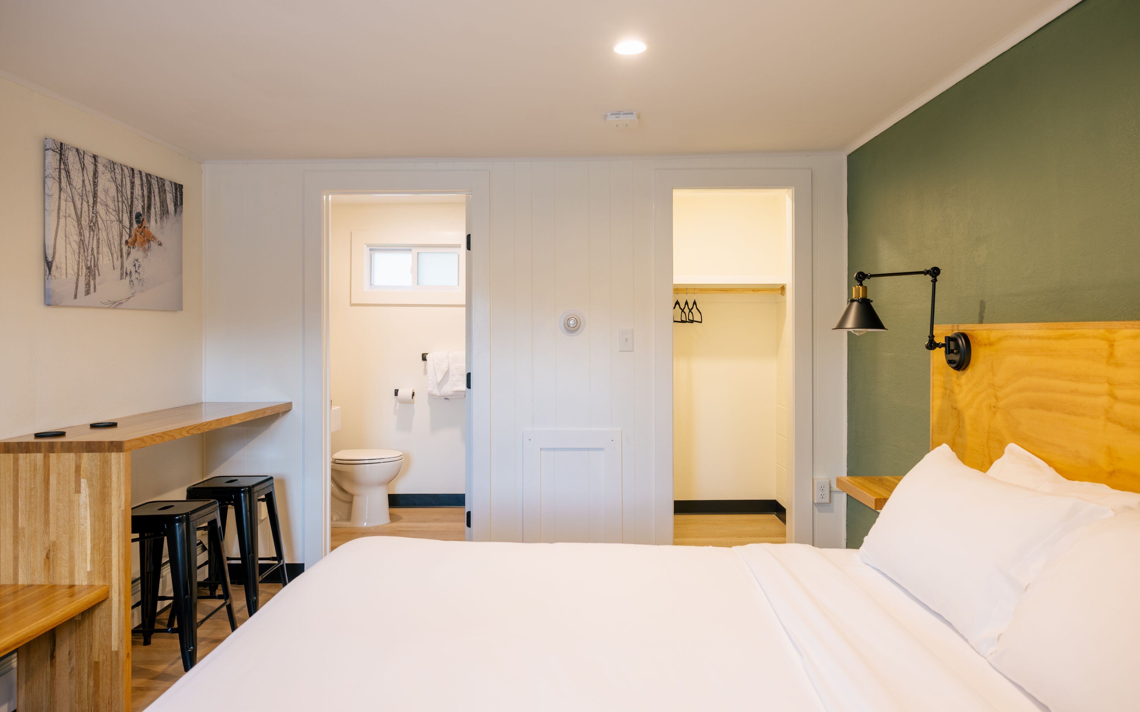 A hotel room with a bed, a desk, a chair, a wooden headboard, a nightstand, a lamp, a painting, and a bathroom visible through an open door.