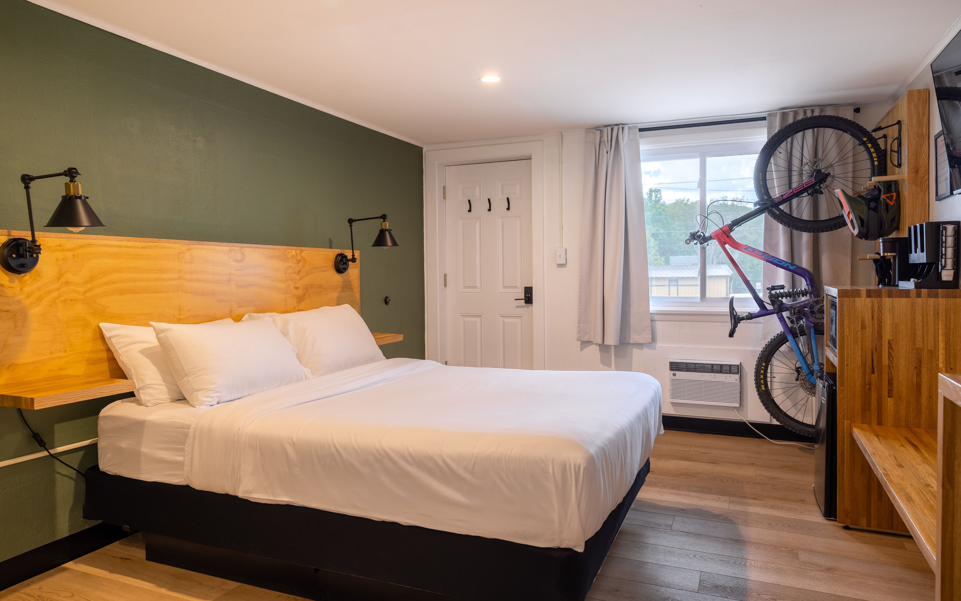 Une chambre avec une tête de lit en bois, un lit recouvert de draps blancs, un vélo accroché au mur et une fenêtre avec des rideaux.
