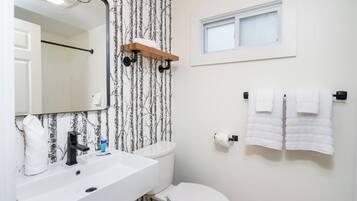 A bathroom with a white sink, black faucet, and a mirror above it. There is a window above the toilet and a towel rack with towels.