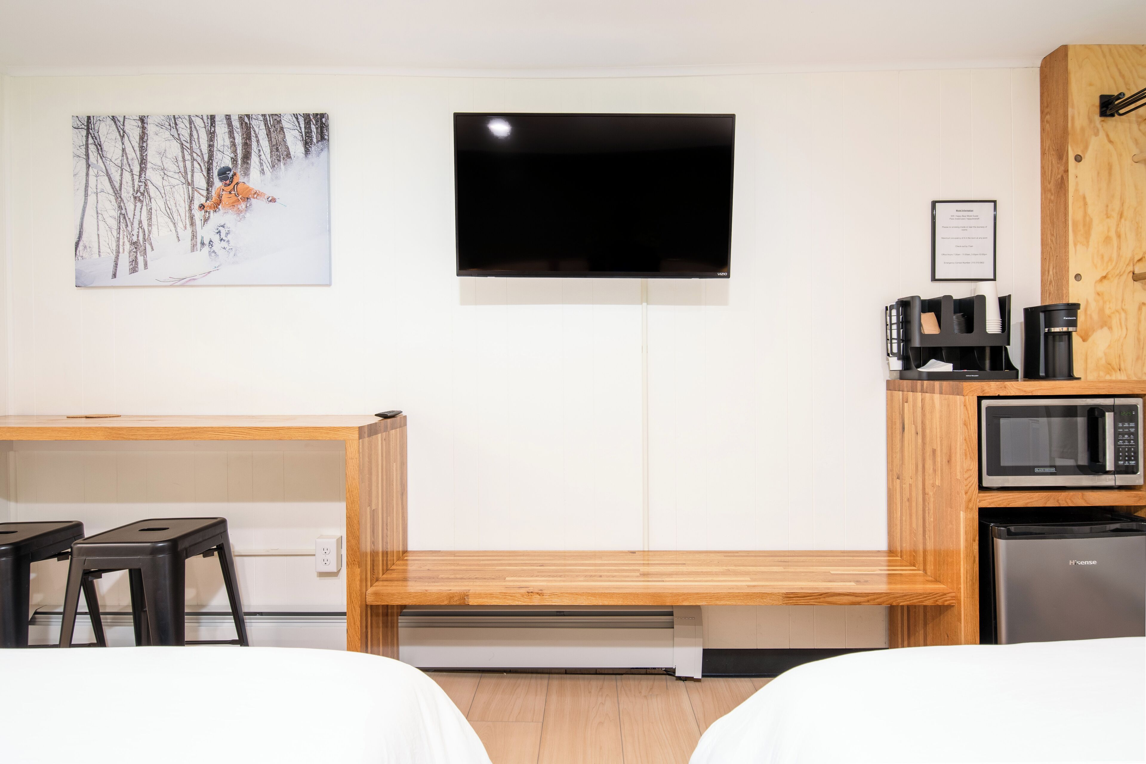 Habitación de hotel con cama, escritorio con dos taburetes negros, televisor montado en la pared, microondas y refrigerador.