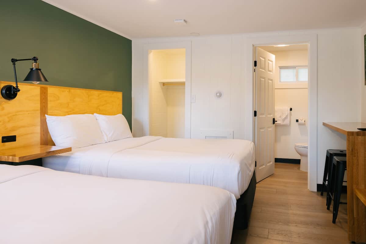 A hotel room with two beds, a wooden headboard, a desk with a lamp, and a bathroom visible through an open door.