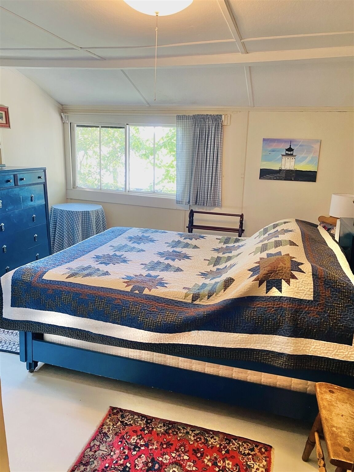 A bedroom with a bed, a dresser, a window with curtains, a painting, and a rug.