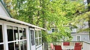 Una terraza de madera con asientos exteriores, un espacio cubierto con ventanas y vista a una calle arbolada.