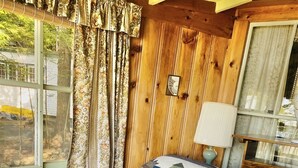 Interior de una cabaña de madera con una cama, ventana, cortinas y una lámpara.