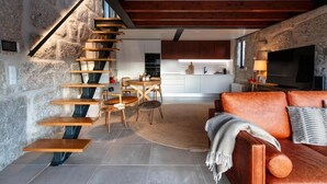 A modern interior with a stone wall, wooden staircase, a cozy seating area with a sofa and chairs, and a kitchen in the background. - Javardo Family House  (Marco de Canaveses)