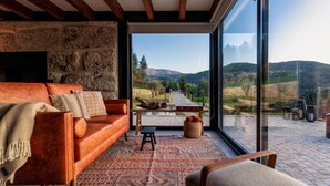 A modern living room with a stone wall, a leather sofa, and a large glass door leading to a scenic outdoor view. - Javardo Family House  (Marco de Canaveses)