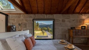 A bedroom with a stone wall, a bed with white and orange pillows, a wooden table with hats, and a view of a garden and mountains through a window. - Javardo Family House  (Marco de Canaveses)