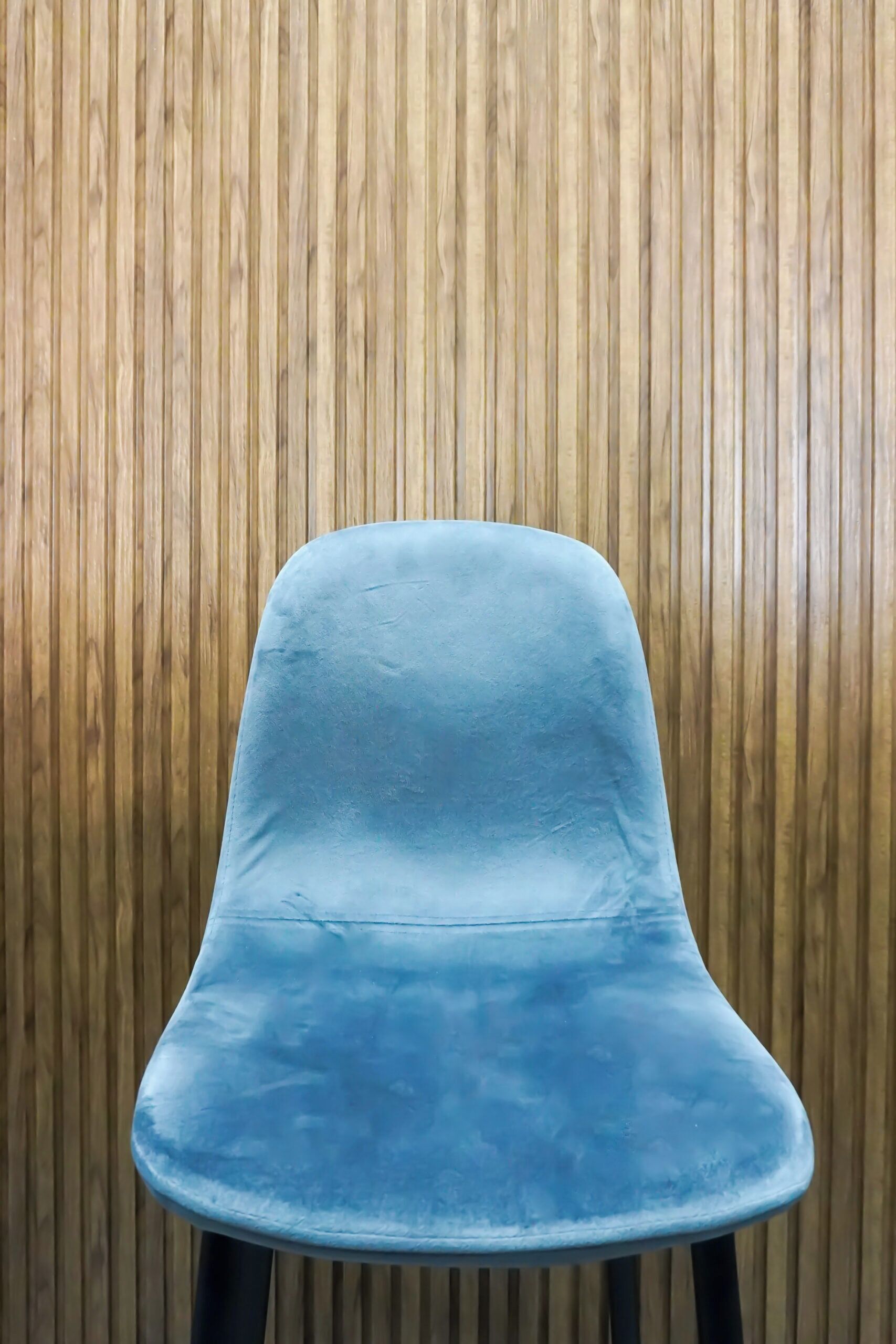 A blue chair with a curved backrest and black legs, positioned against a wooden wall.