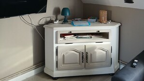 A room with a TV mounted on the wall, a white cabinet with drawers, a lamp, and a remote control on a black couch.
