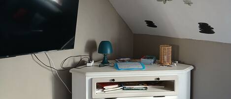 Une pièce avec un téléviseur fixé au mur, un meuble blanc avec des tiroirs, une lampe et une télécommande sur un canapé noir.
