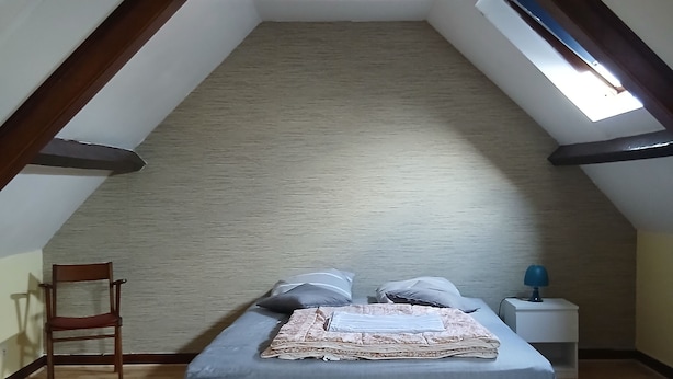 A bedroom with a sloped ceiling, a bed with pillows, a chair, and a nightstand with a lamp.