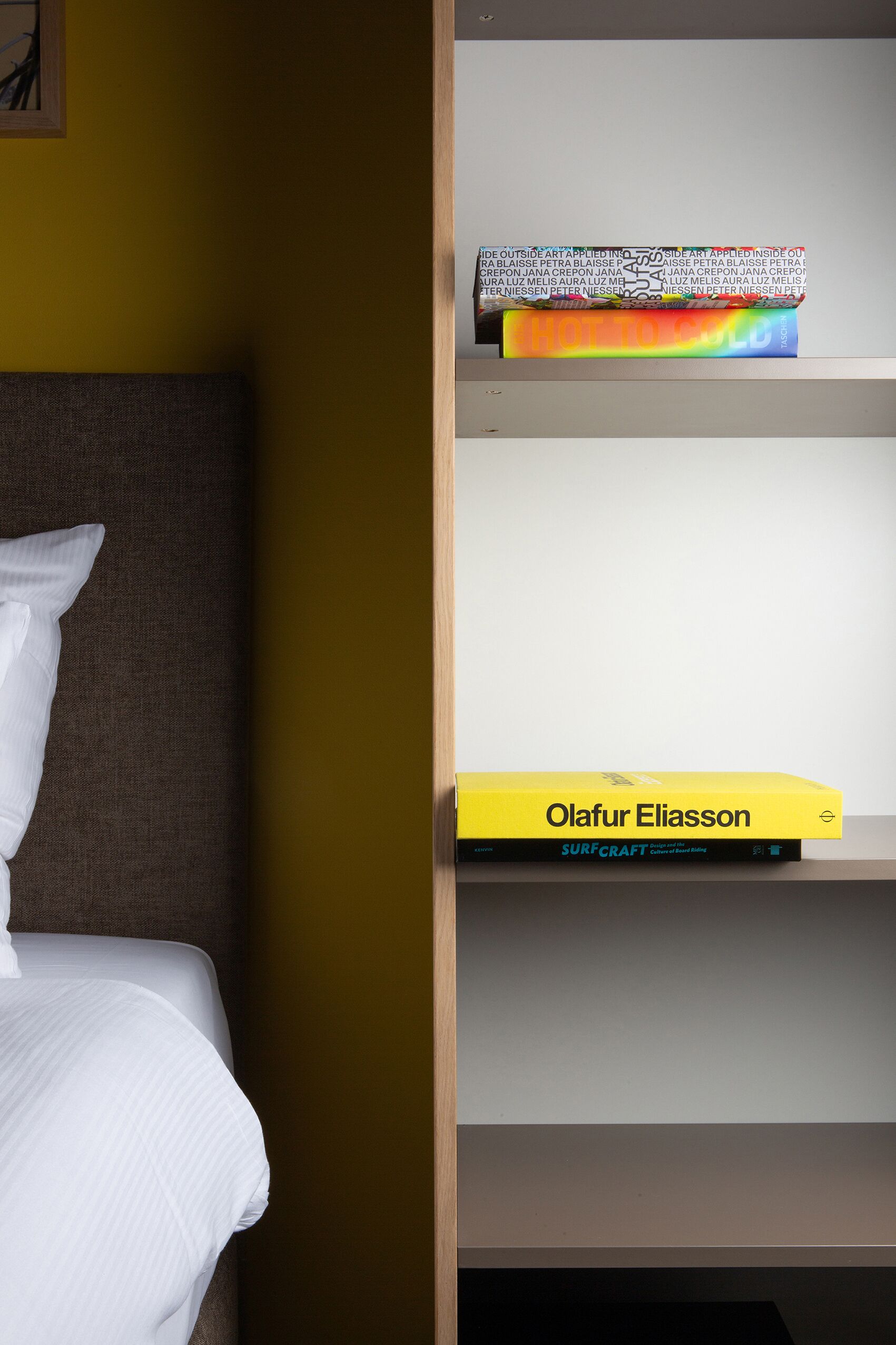 A hotel room with a bed, a bookshelf, and a yellow book titled 'Olafur Eliasson'.