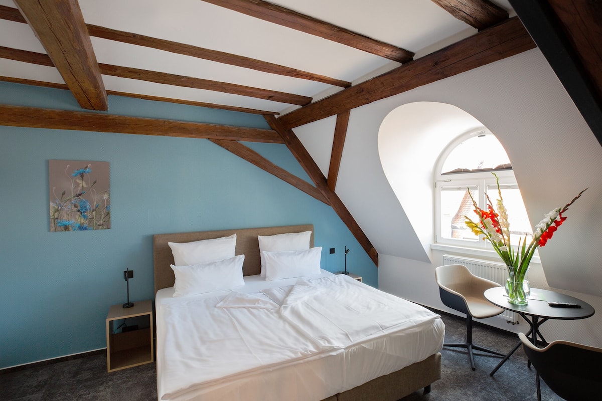 A bedroom with a large bed, a nightstand, a chair, a small table with a vase of flowers, and a window with a view of the outside.