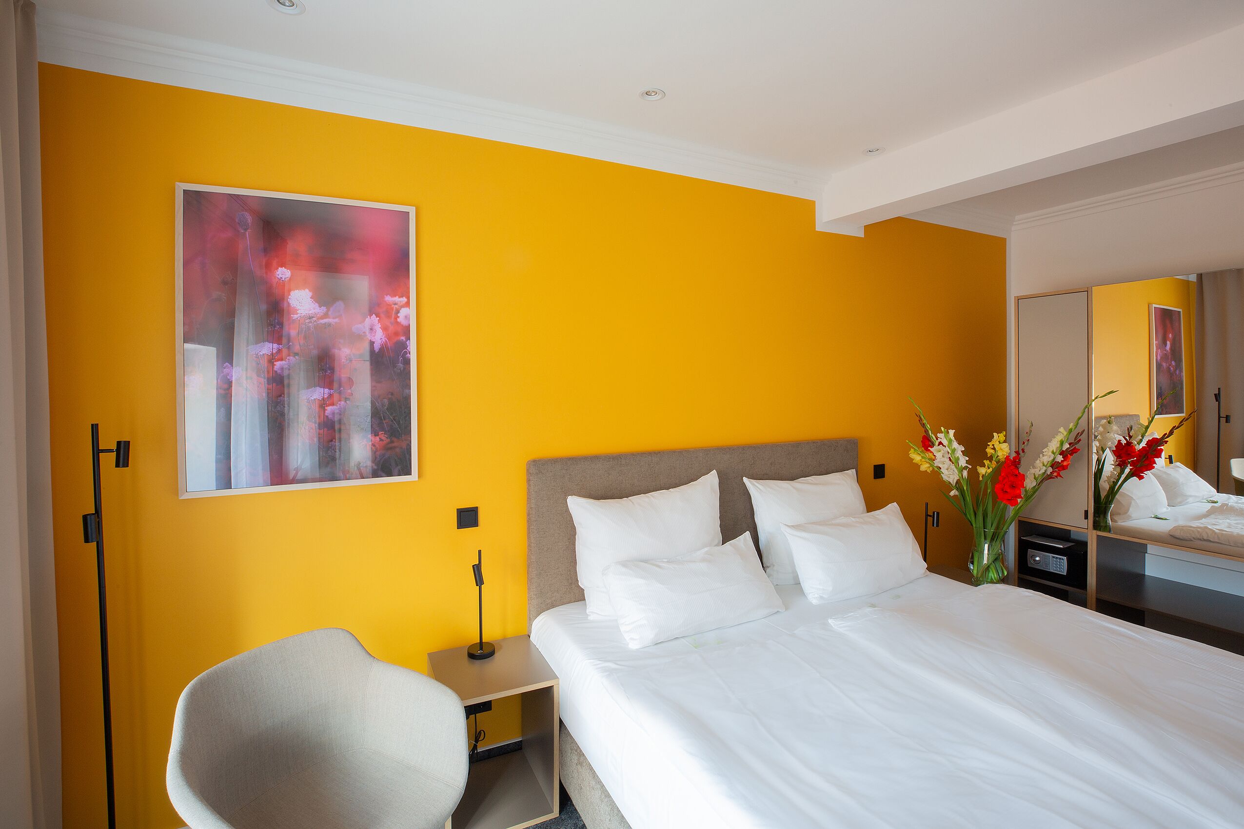 A hotel room with a large bed, a bedside table, a chair, a nightstand, a mirror, and a painting on the wall.