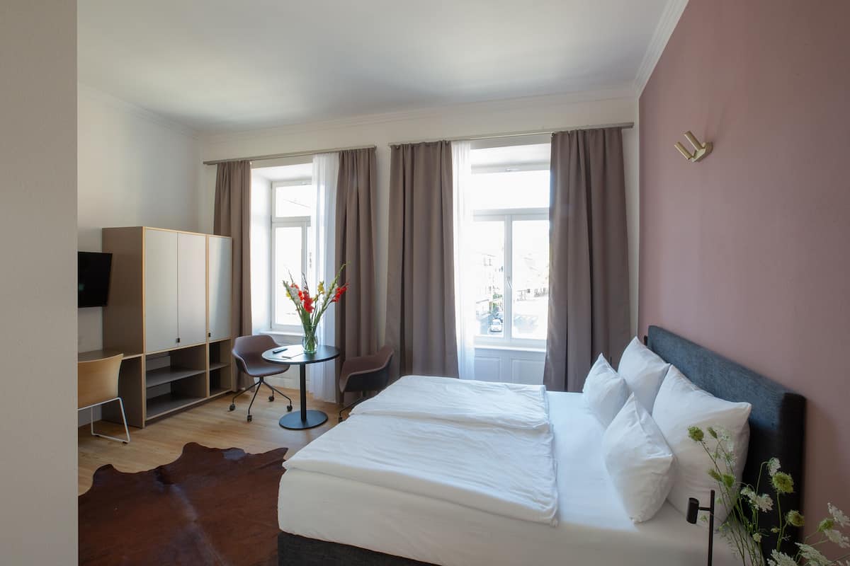 A hotel room with a bed, a desk, a chair, a TV, and a window with curtains.