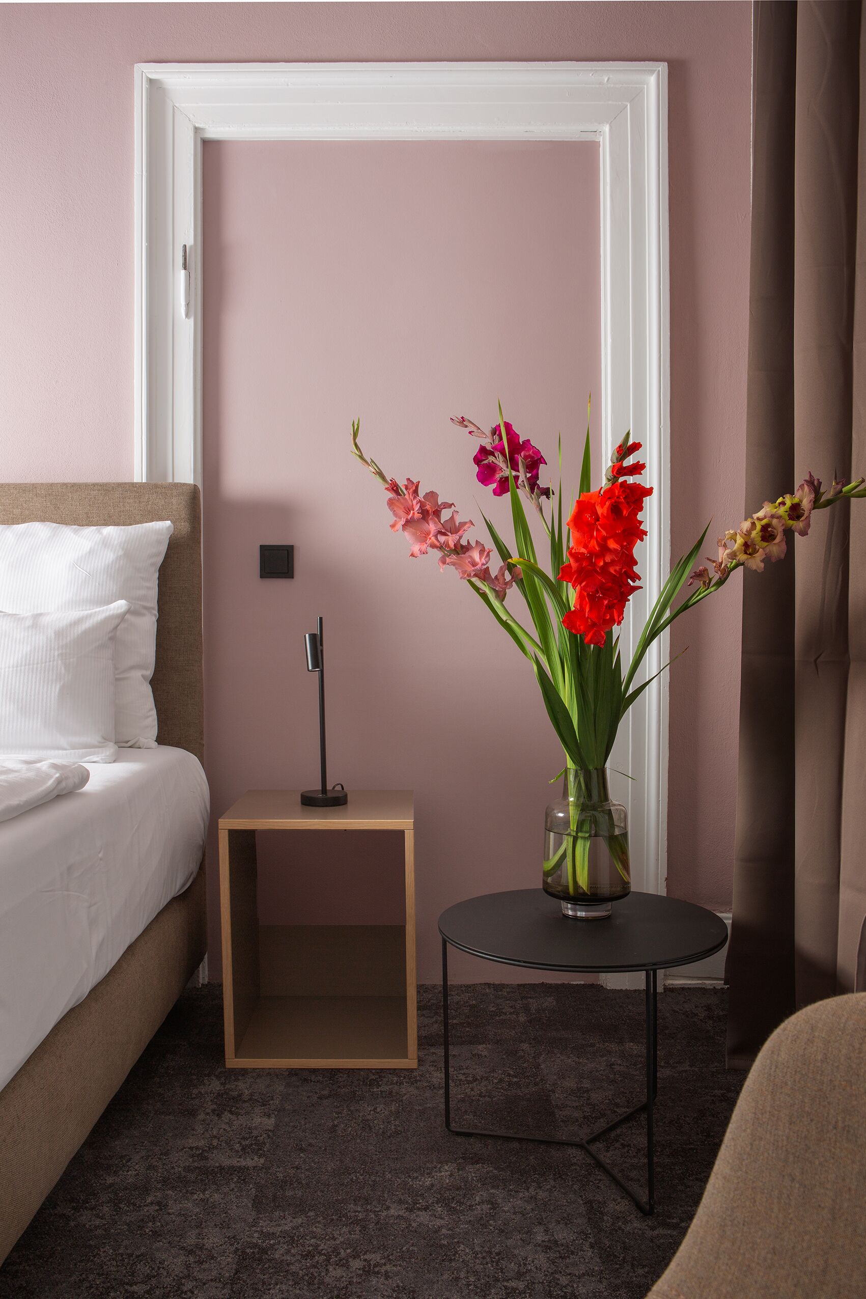 A neatly arranged bedroom with a bed, a bedside table with a lamp, and a vase of flowers.