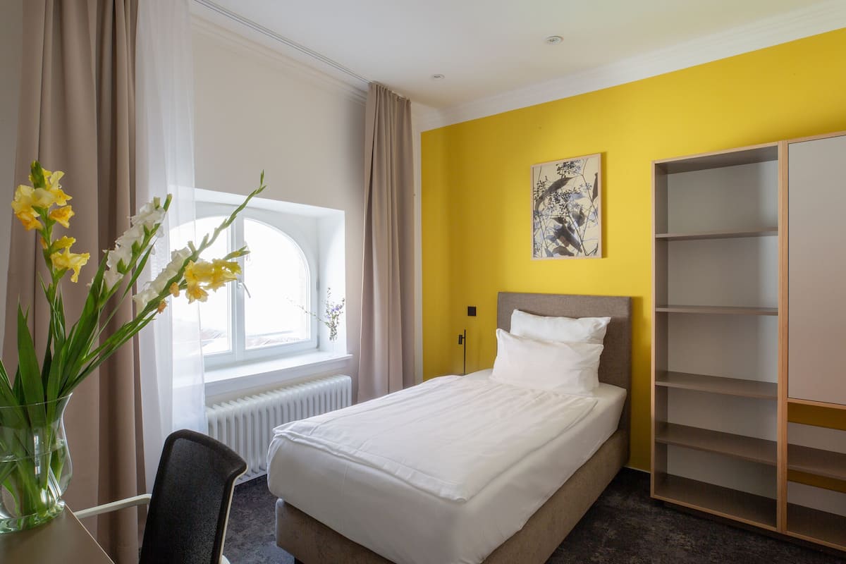 A hotel room with a bed, a window with curtains, a vase with flowers, and a wall with a picture.