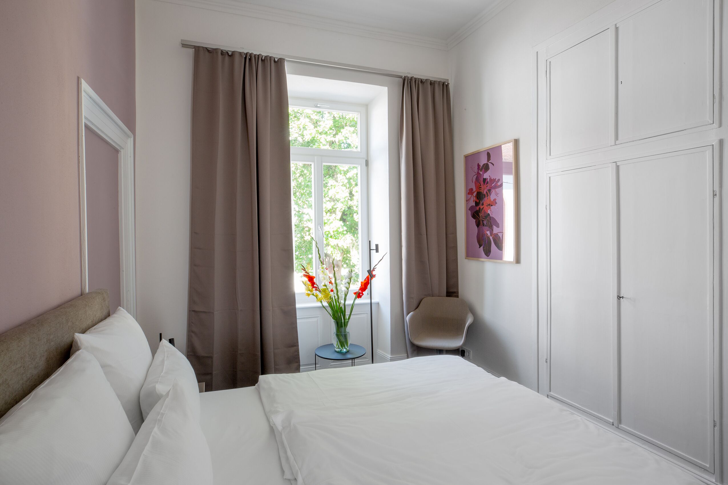 A bedroom with a bed, a chair, a window with curtains, a vase with flowers, and a door.