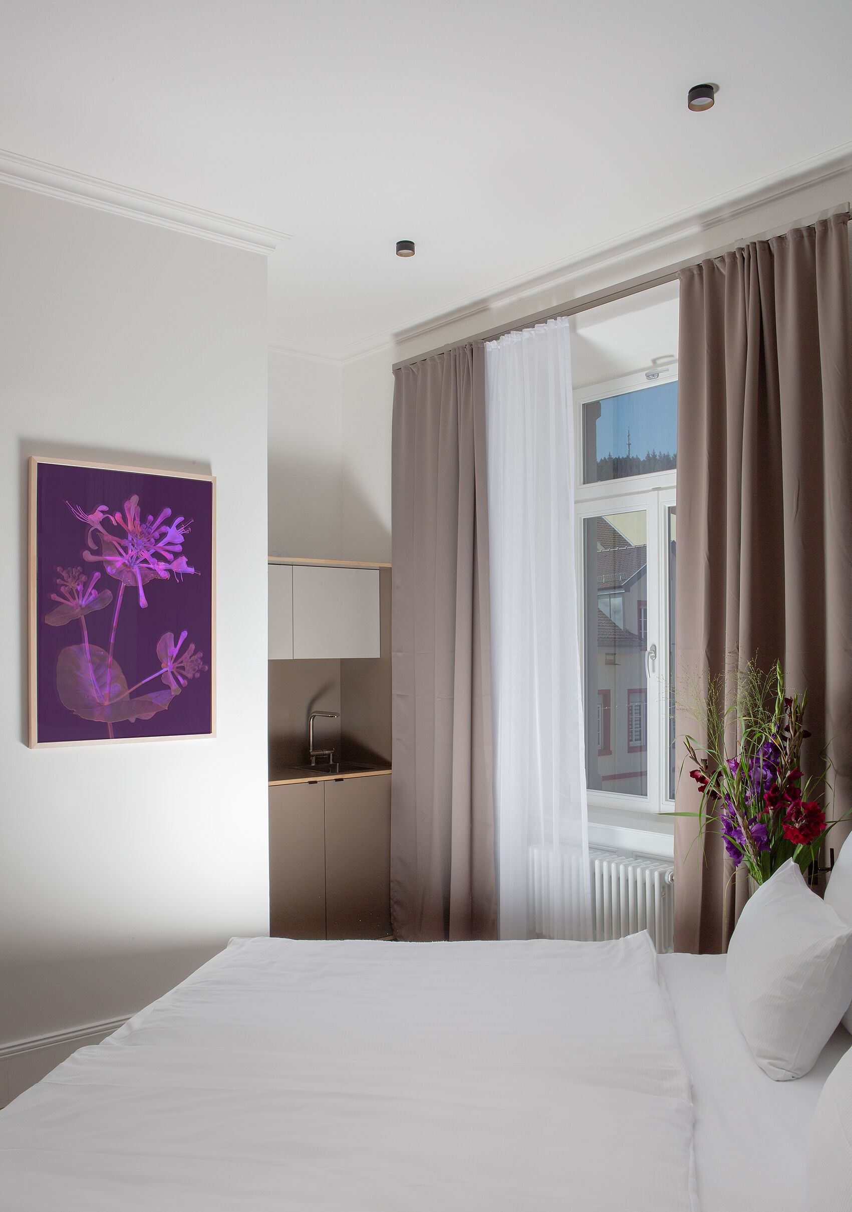 Een hotelkamer met een bed, een schilderij, een raam met gordijnen, een radiator en een kleine kitchenette.