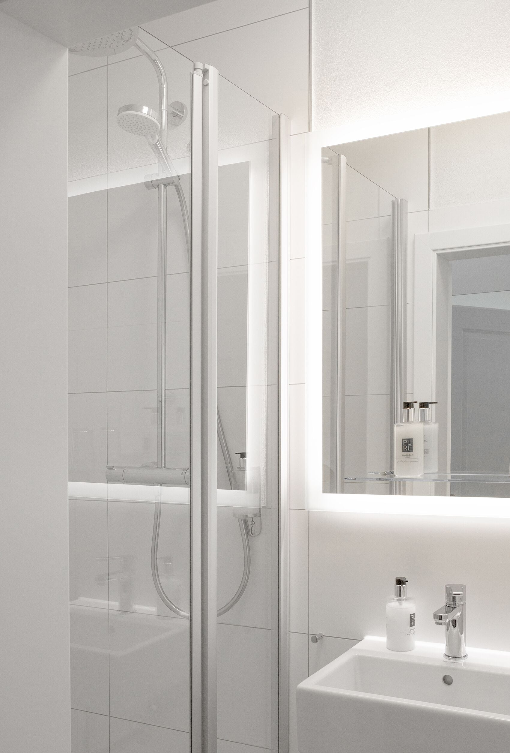 A modern bathroom with a glass shower enclosure, a sink with a mirror above it, and a bottle of Pure Essence on the sink.