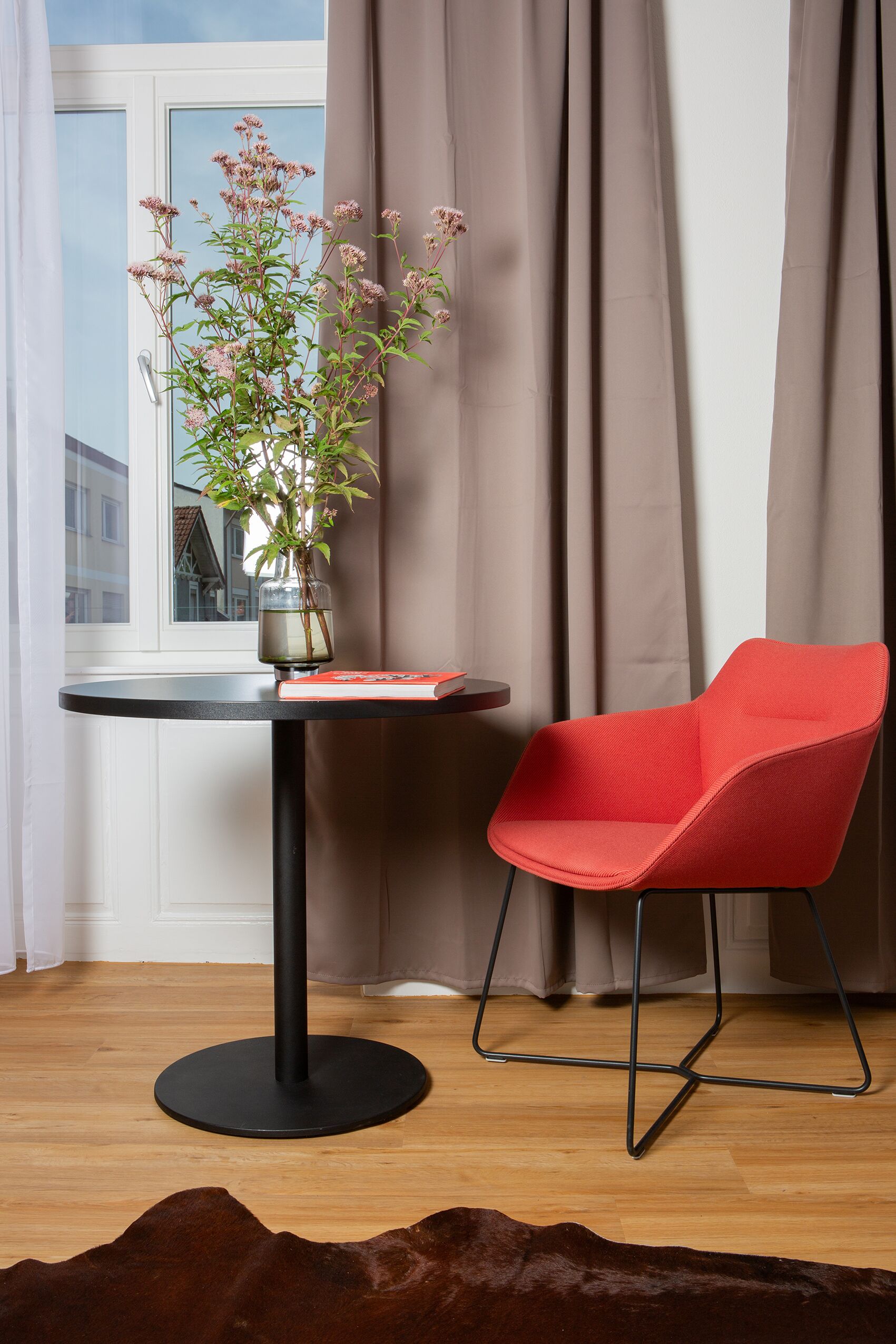Um quarto com uma mesa redonda preta, uma cadeira vermelha, um vaso com flores e uma janela com cortinas.