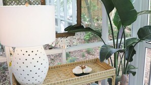 A wicker bench with rolled towels, a white lamp, and a mirror with a floral frame in a sunlit room with large windows and green plants. - North End, 3 Br Luxury Cottage, Pet Friendly, Private Road, 5 min walk to Beach (Tybee Island)