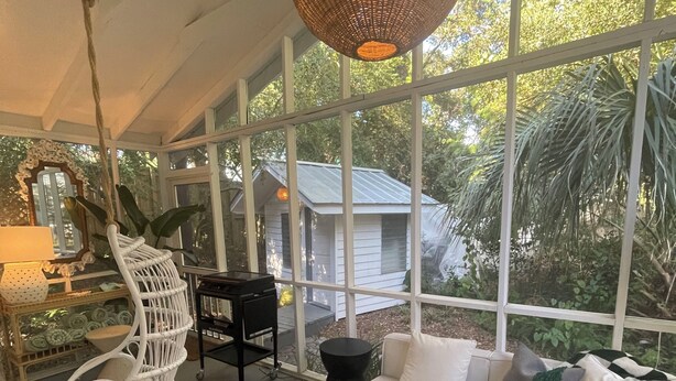 A sunlit room with a white sofa, black round tables, a wicker hanging light fixture, and a view of greenery outside. - North End, 3 Br Luxury Cottage, Pet Friendly, Private Road, 5 min walk to Beach (Tybee Island)