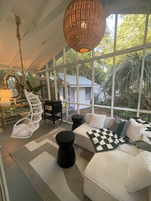 Une pièce baignée de lumière, avec un canapé blanc, des tables rondes noires, un luminaire suspendu en osier et une vue sur la verdure à l’extérieur.