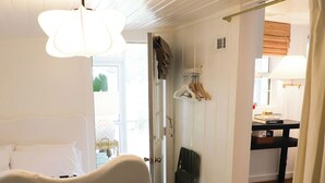 A cozy room with a white armchair, a striped stool, and a small table. There is a door leading to a kitchen area with a wooden floor. - North End, 3 Br Luxury Cottage, Pet Friendly, Private Road, 5 min walk to Beach (Tybee Island)