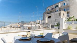 Terraza en la azotea con mesas listas para comer, con vistas a un paisaje urbano costero con edificios y cielo despejado.